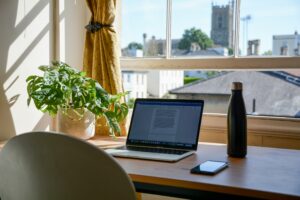 a macbook, water bottle, phone, and plant on a table that is in front of an open window