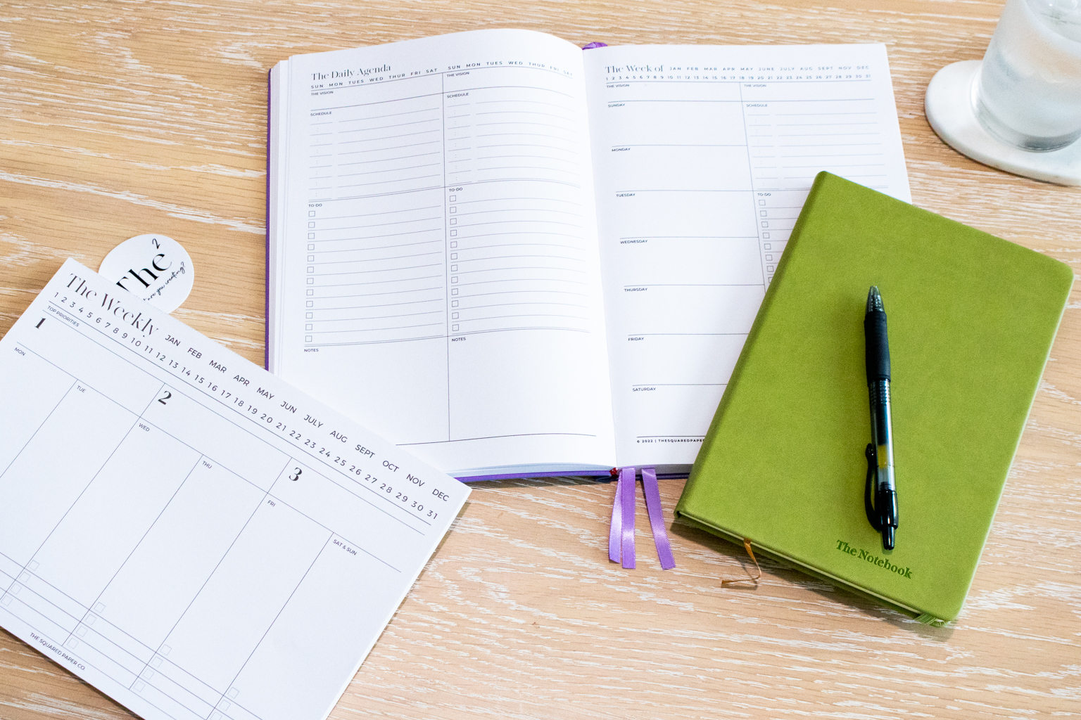 A lilac copy of The Square Paper Co’s Undated Planner laying open on a wooden desk. A weekly desk planner insert is also pictured to the left and an olive coloured notebook with a pen is pictured to the right.
