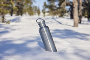 Stainless steel water bottle, which fits the equivalent of a bottle of wine, stuck into the snow with a picturesque winter landscape behind.