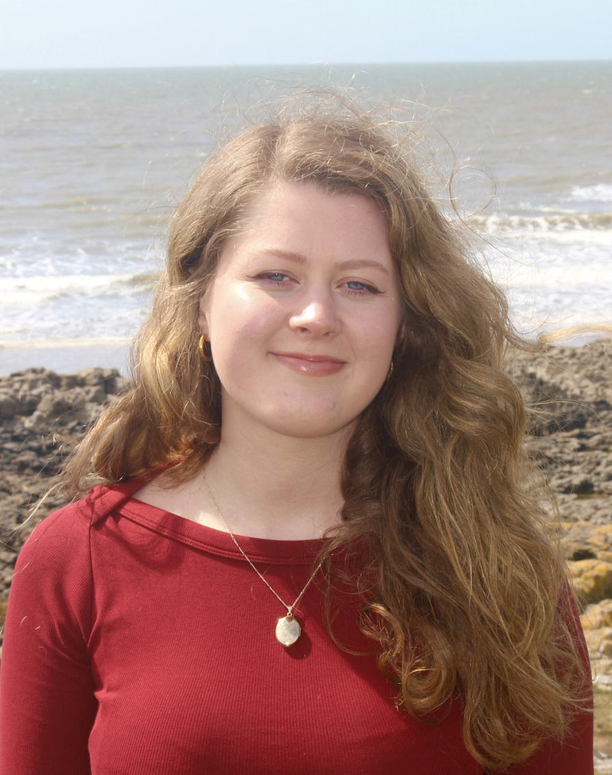 Amelia Jones, a white woman with blonde hair, smiles to camera in front of the sea. She wears a red top with a gold locket around her neck.
