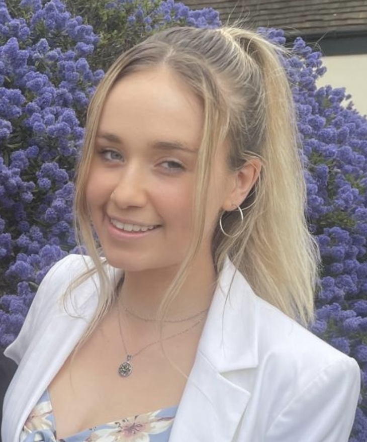 Izzy, a white woman with blonde hair, stands in front of purple flowers, looking to camera
