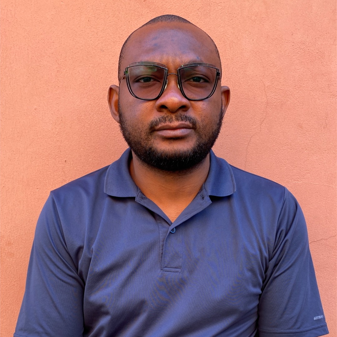 Vining Ogu, a Black man wearing glasses and with a trimmed beard, looks to camera. He wears a blue shirt.